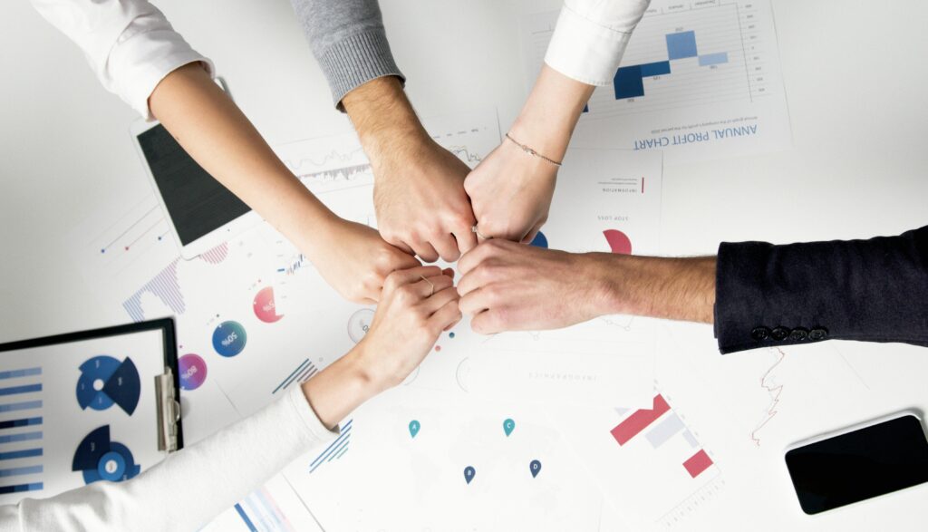 Five people's hands, palm-down, reaching toward each other in cooperation.